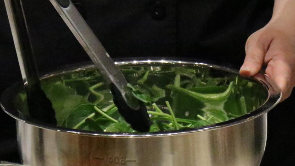 A pair of hands use tongs to mix up a bowl of spinach salad.