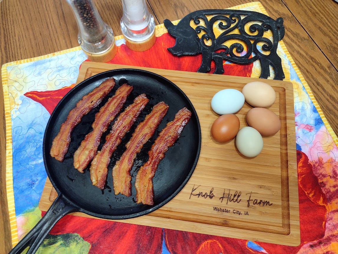 Cooked bacon on a cast iron skillet, set on a Knob Hill Farm wooden cutting board next to colorful eggs