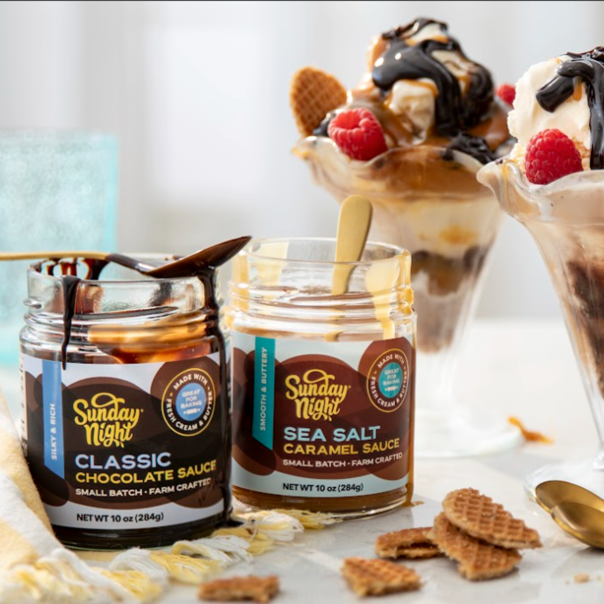 Jars of Sunday Night Foods Chocolate Sauce and Caramel sauce are opened with spoons ready to use. Two sundaes of vanilla ice cream are covered in the sauces, raspberries and waffle cone pieces.
