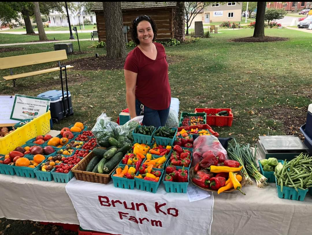 A woman in a red shirt stands behind tables covered in fresh produce in shades of red, orange, and green.