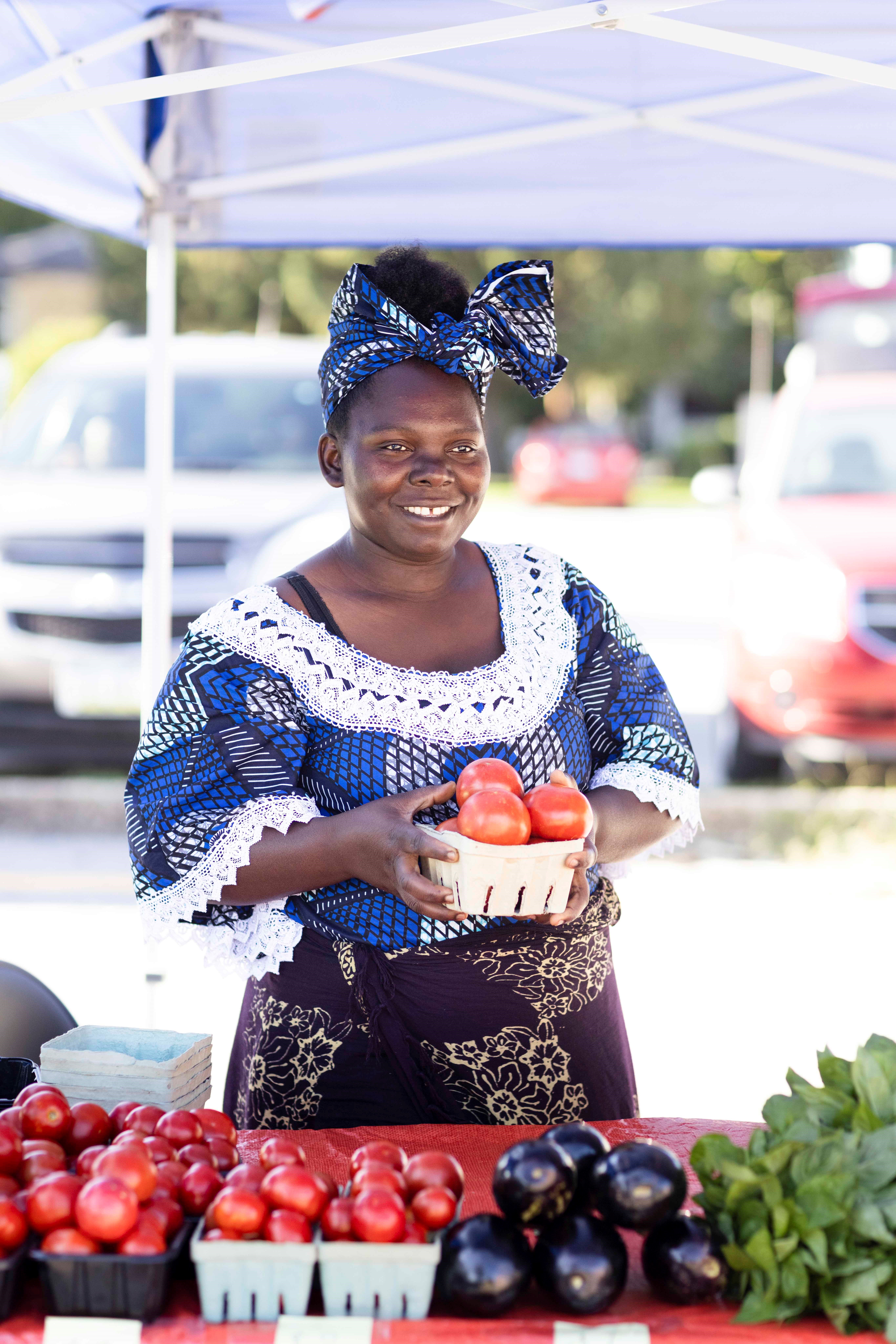 A woman holds a small basket of tomatoes.  She stands behind a table covered in other sorts of fresh produce.