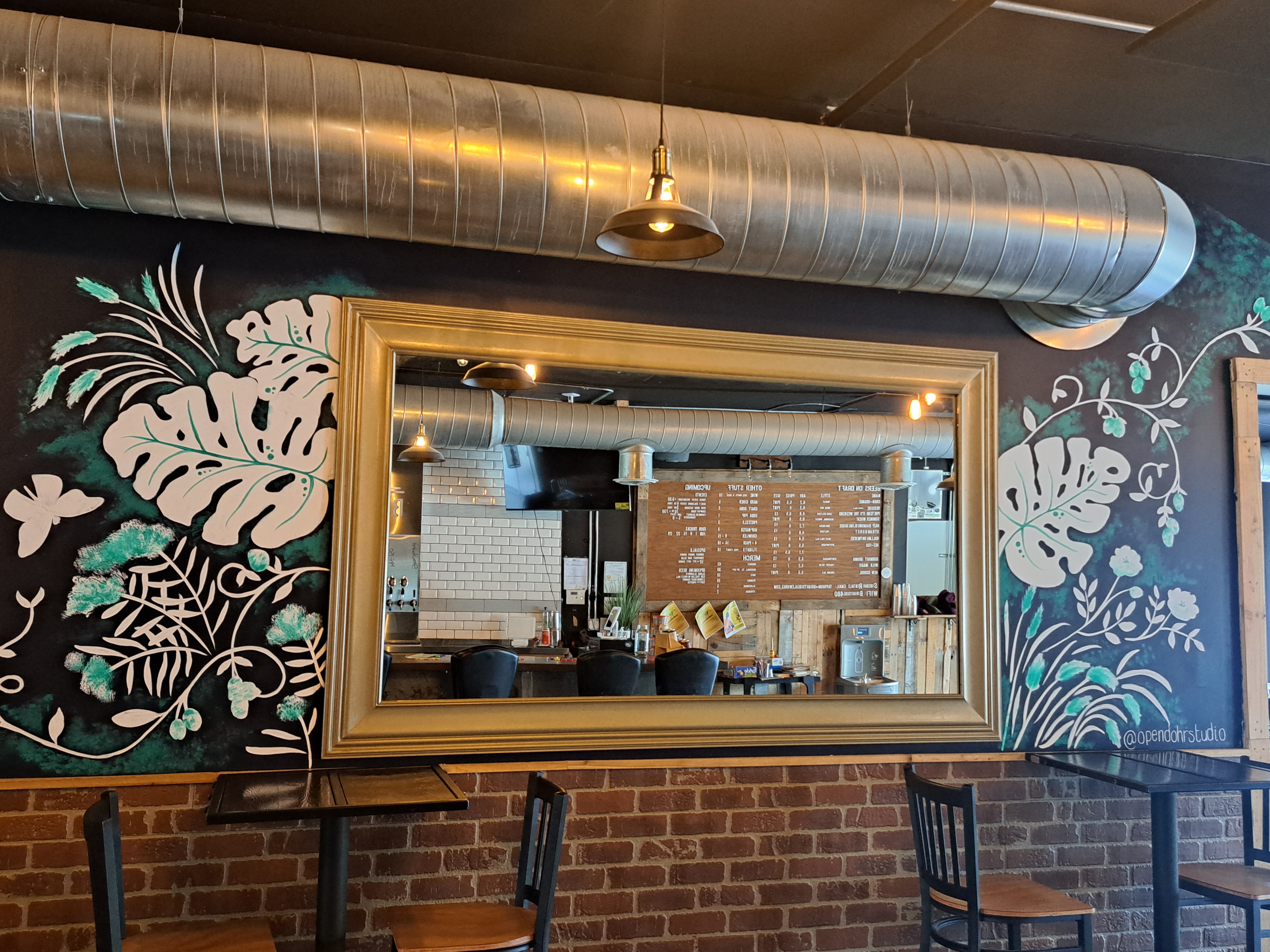 A mirror is surrounded by a mural done in green, white, and black of leaves and other plants.  Tables with chairs are under the mirror.  This is inside Brightside Aleworks.