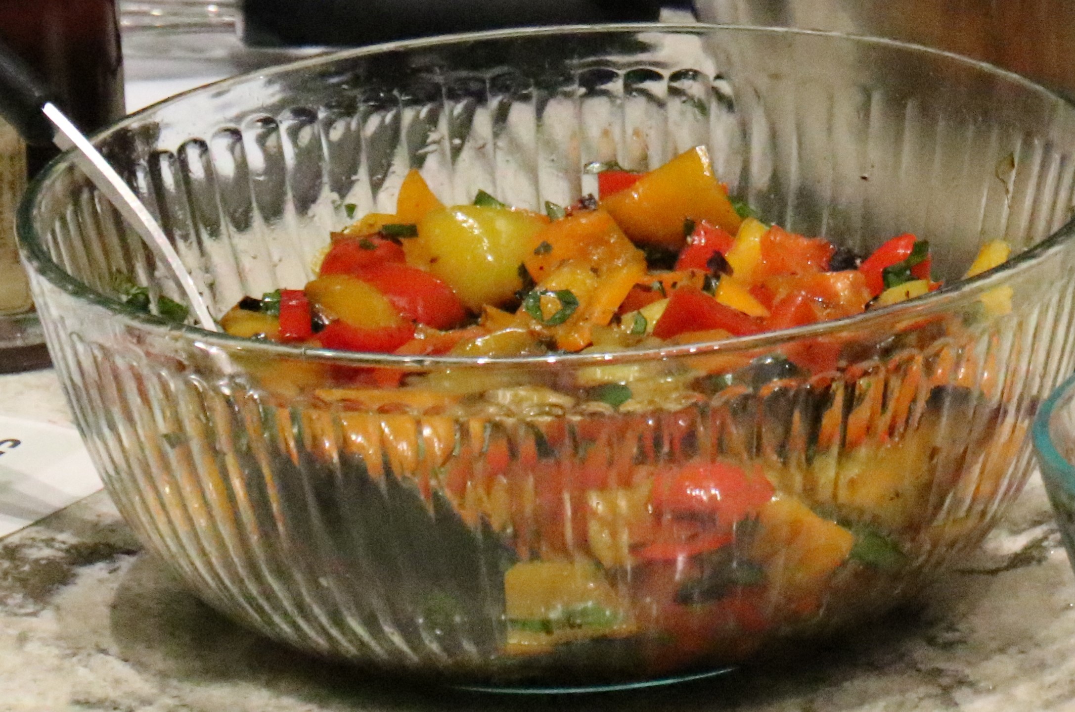 A clear bowl holds a bunch of grilled peppers with basil and olive oil.