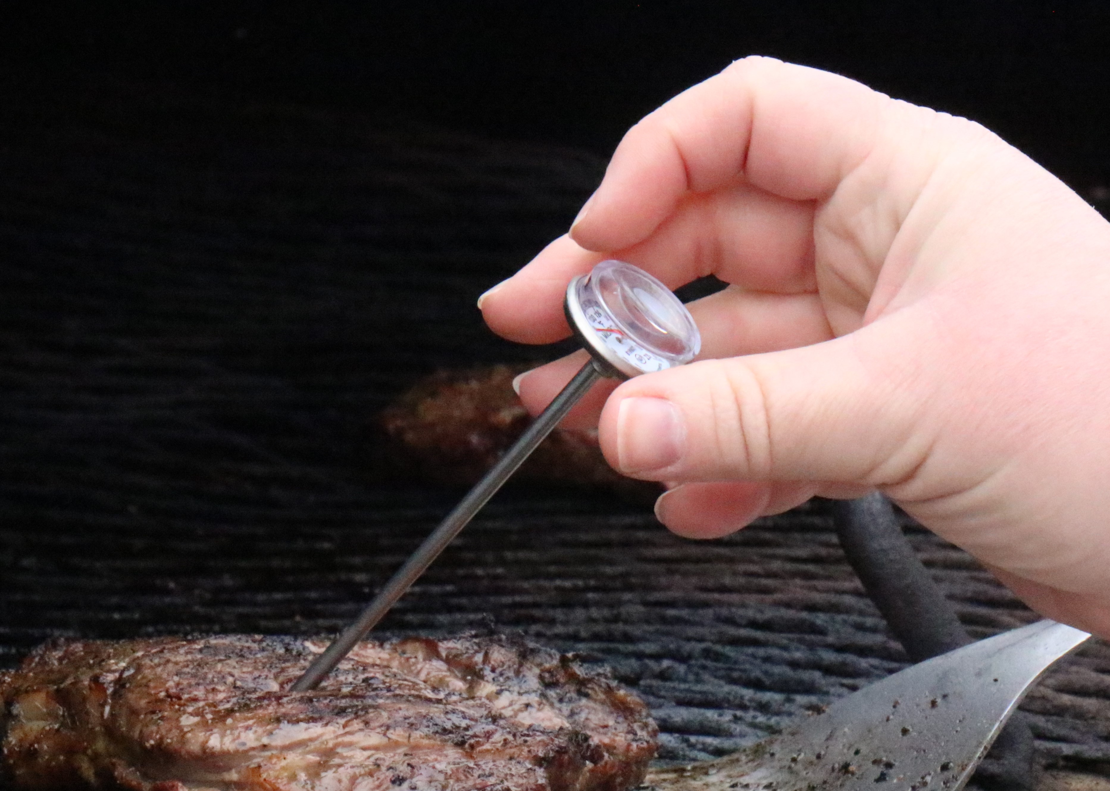 A hand with a thermometer checks the temperature of a steak on a grill.