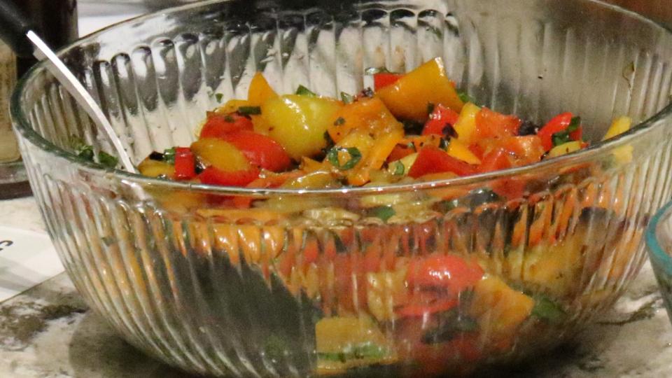 A clear bowl holds a bunch of grilled peppers with basil and olive oil.