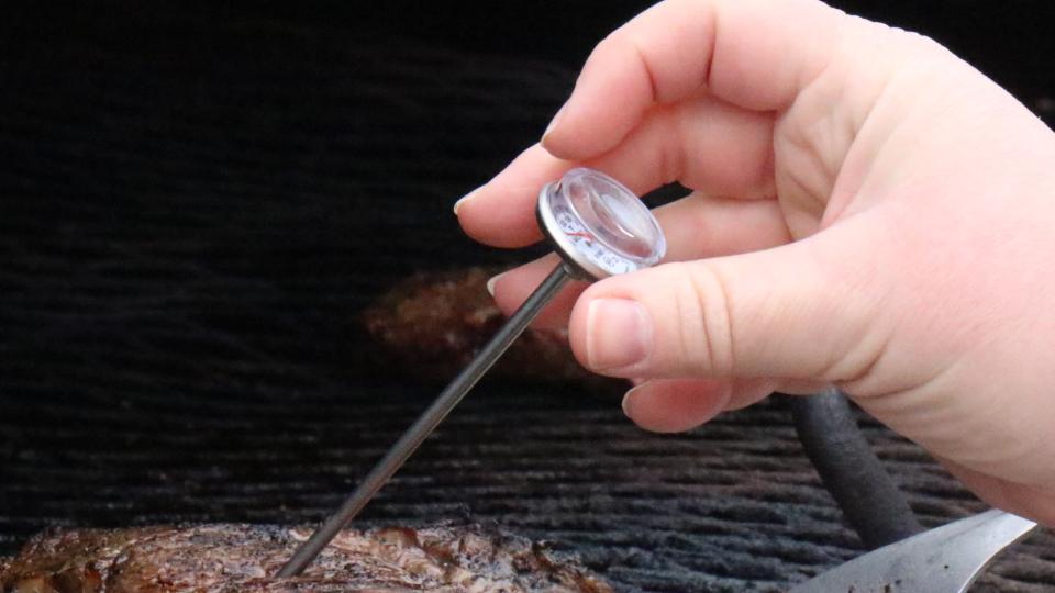 A hand with a thermometer checks the temperature of a steak on a grill.