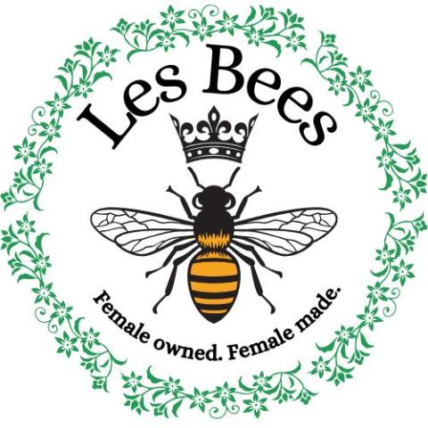 In the center of the Les Bees logo is a bee with a crown and the Les Bees name above its head and the phrase "Female owned. Female made." below the bee. All of this is inside a green circular border of leaves and flowers.