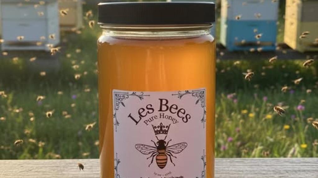 Jar of honey from Les Bees on a wood ledge with sunlight shining through and bees flying in background.